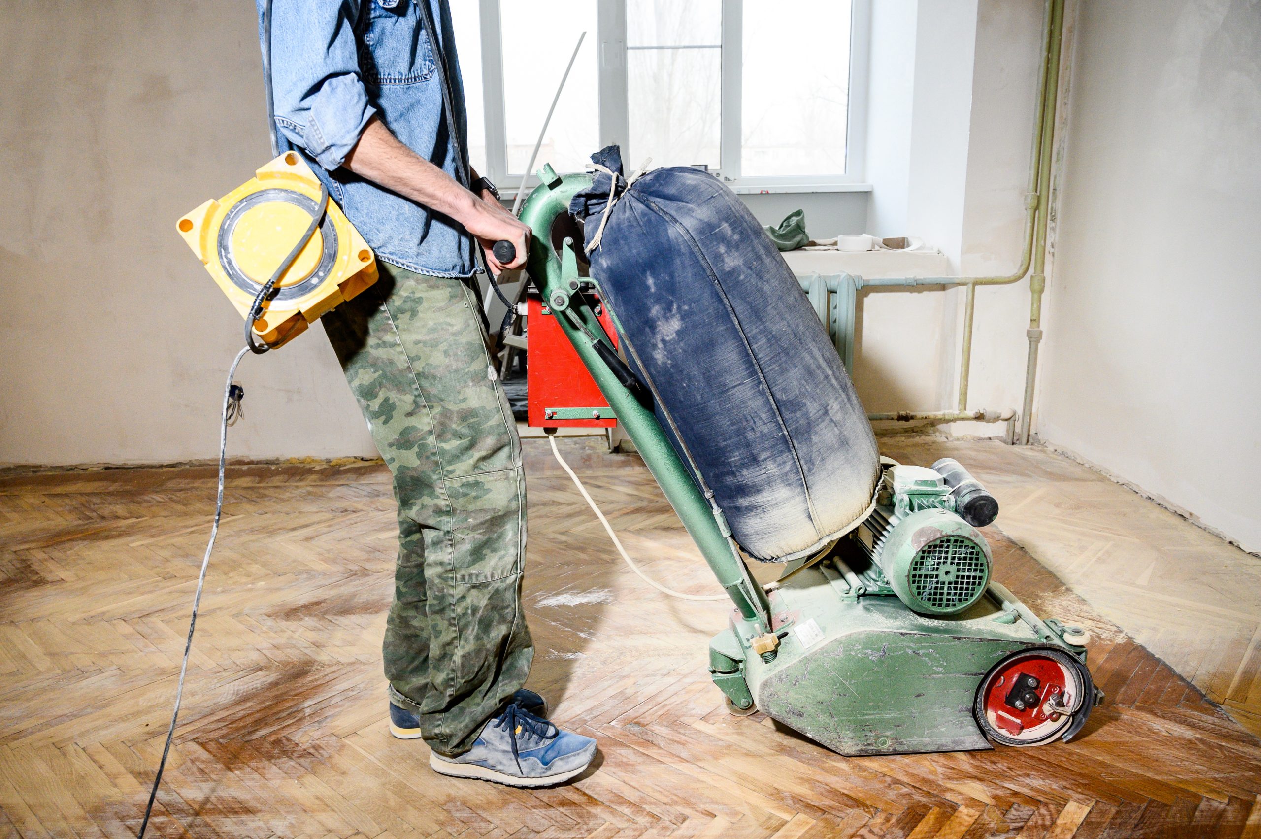 Professional wood floor sanding in Greenwich being carried out by a technician with a drum sander on a worn herringbone parquet floor.