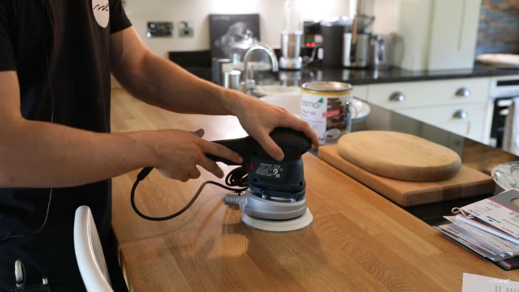 Kitchen Worktop Sanding in Greenwich – professional using a random-orbital sander on a solid oak worktop in a modern kitchen; oak hardwood kitchen floor.