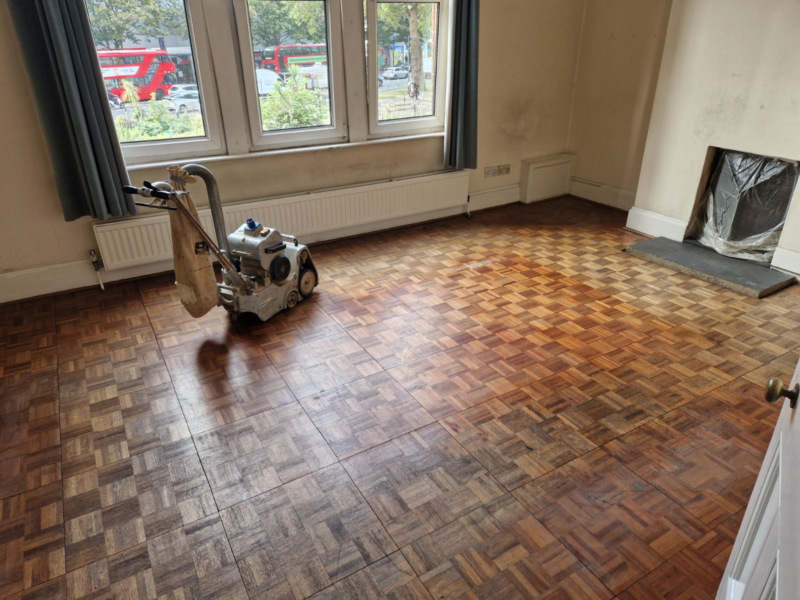 Wood Floor Sanding in Greenwich on an oak mosaic parquet floor, with a sanding machine part-way through restoring a worn living room.